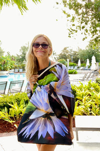 Artist Shelly Lawler holding a colorful lotus -patterned beach bag in an outdoor setting with greenery and a pool.