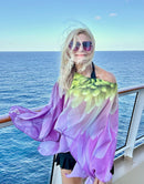 Petals Poncho worn on a cruise deck, showing pink and green dahlia artwork with lightweight fabric flowing in the wind against an ocean backdrop