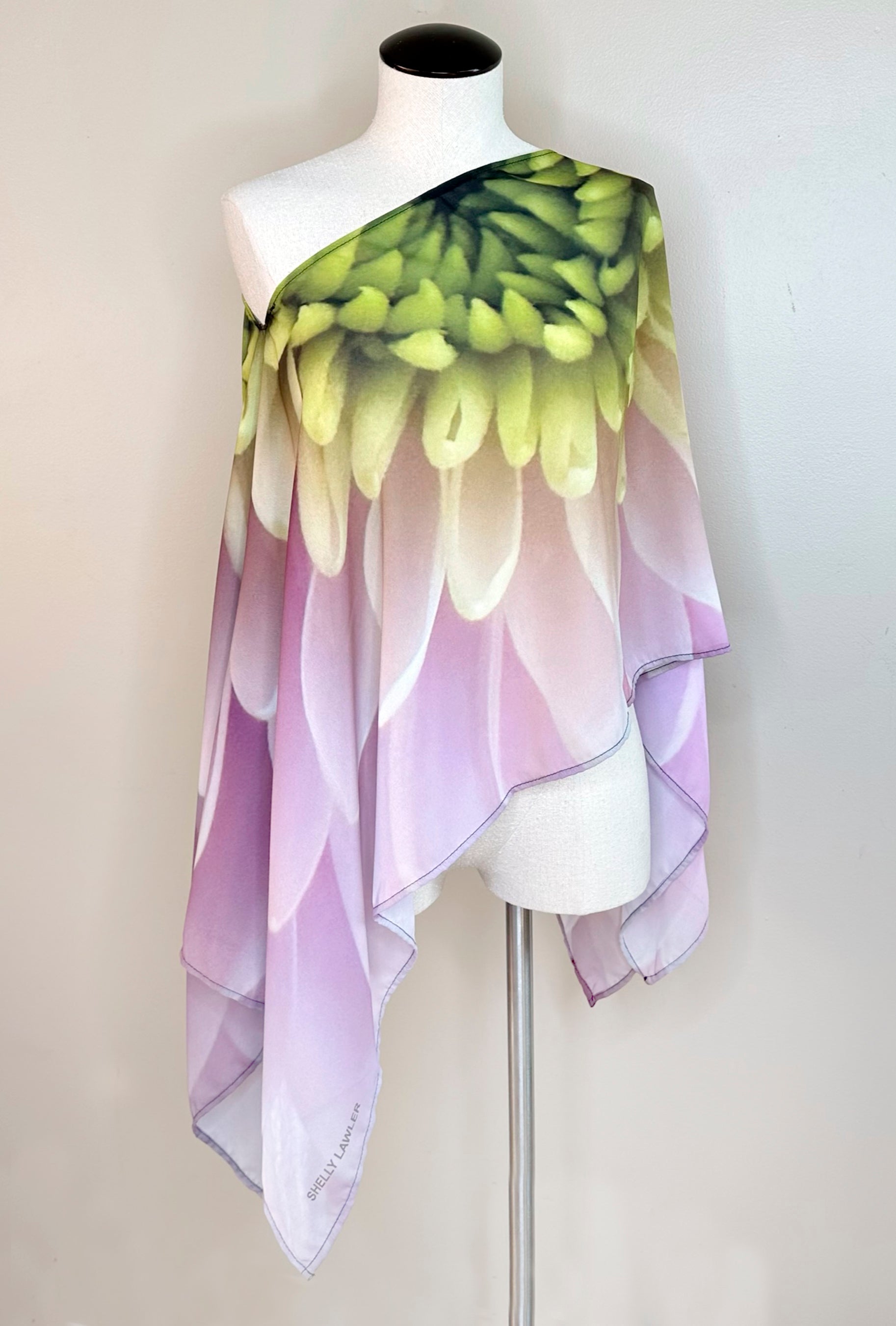 Petals Poncho displayed on a mannequin, draped off one shoulder showing asymmetrical styling with pink and green dahlia artwork and lightweight flowing fabric