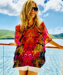 Red Poncho worn as a tunic, showing autumn-toned abstract artwork with lightweight fabric flowing in the wind against an ocean backdrop
