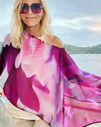 Royal Poncho draped off one shoulder, showing abstract floral artwork in purple, pink, and white with lightweight fabric flowing in the wind against an ocean backdrop